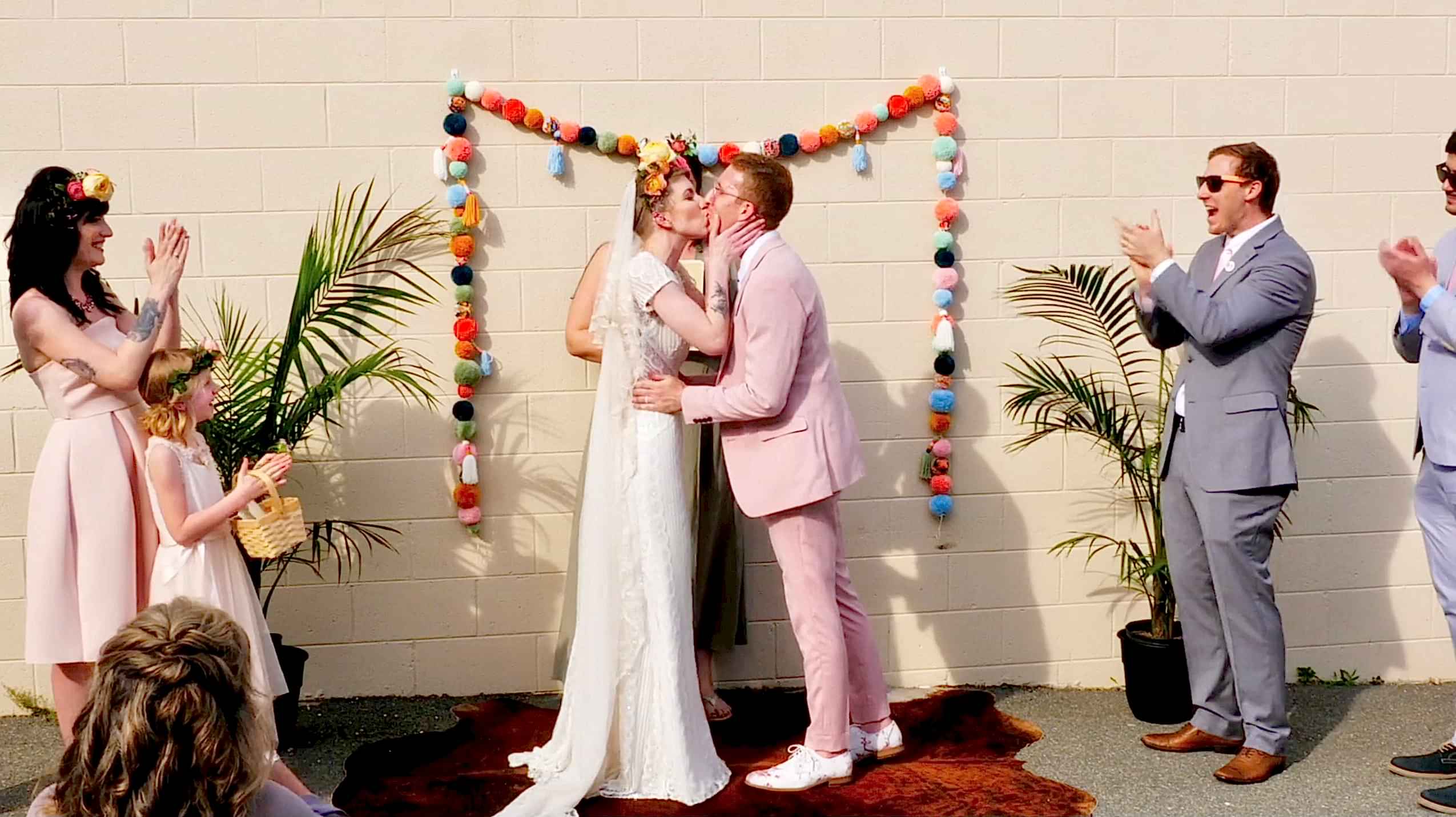 Picture of a couple sharing their first kiss at their wedding. Surrounded by brightly colored decorations - the bride is in a wedding dress and the groom is in a pink suit.