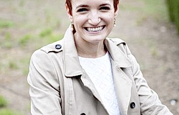 A woman in a white wedding dress and khaki trench coat wears her red hair in a pixie cut with floral headpiece and smiles