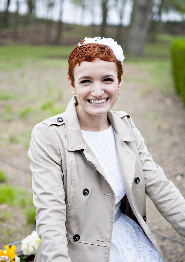 A woman in a white wedding dress and khaki trench coat wears her red hair in a pixie cut with floral headpiece and smiles