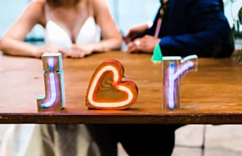 Neon block letters read "j heart r" on a wood table in front of a couple in wedding clothes
