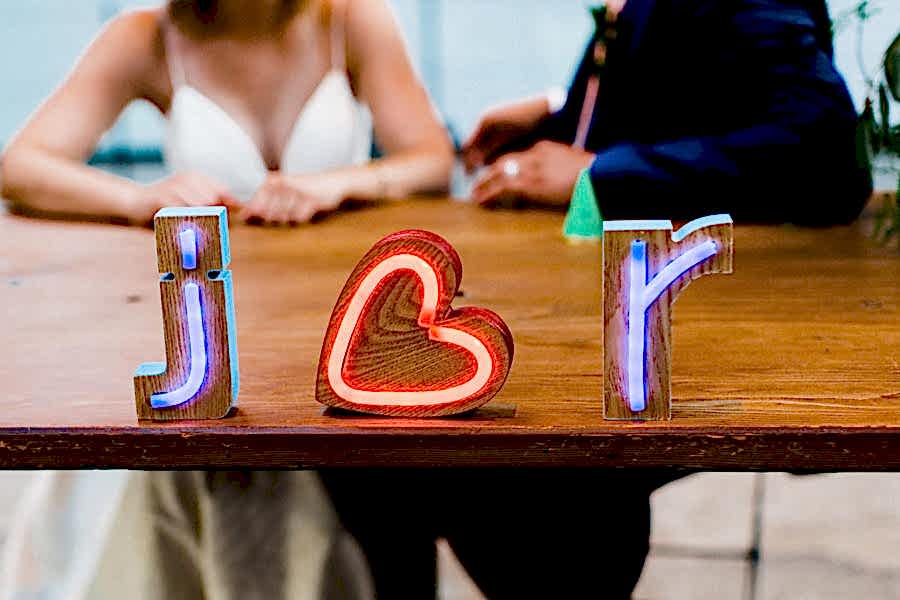 Neon block letters read "j heart r" on a wood table in front of a couple in wedding clothes