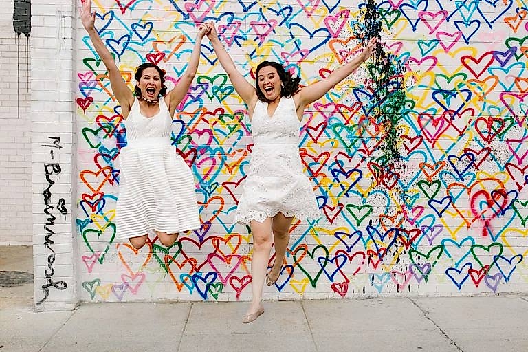 Lesbian couple eloping, jumping in front of a heart wall