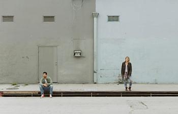 A couple on the street—one sits on the curb and one stands far away from each other, looking in different directions