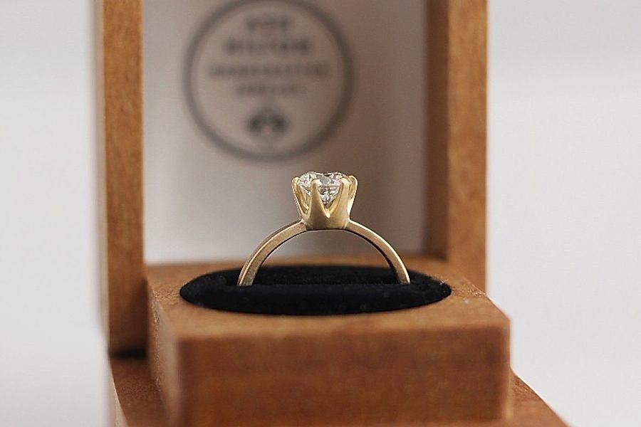 Solitaire diamond engagement ring with silver-colored band from Ash Hilton displayed in a wood ring box