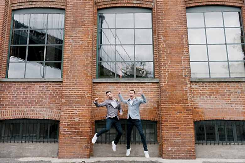 two grooms jumping for joy in front of brick industrial building