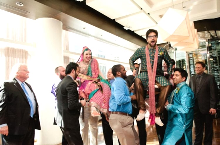 Bride and groom in Indian wedding attire, being lifted in chairs by wedding guests for the Hora