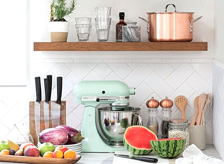 Kitchen counter still life, with copper pan, KitchenAid mixer, knife block, tray of fruit, halved watermelon on cutting board, eggplant on stack of plates 