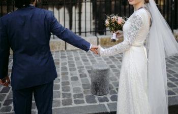 bride in gown with veil, holding bouquet in right hand, holds groom's hand and turns toward you, smiling