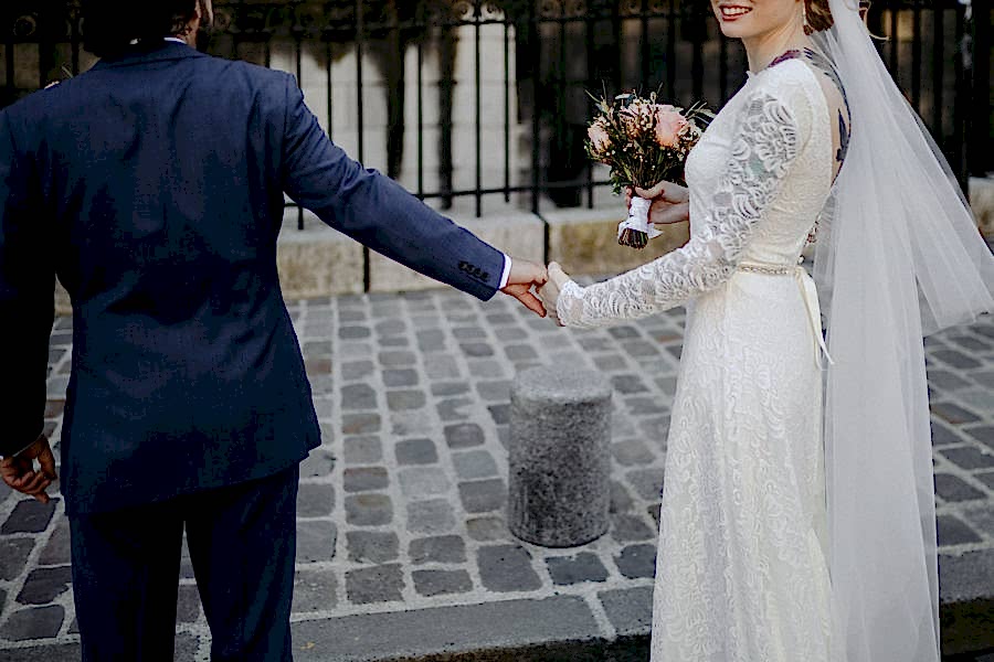 bride in gown with veil, holding bouquet in right hand, holds groom's hand and turns toward you, smiling