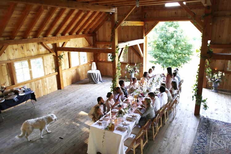 People eating at long family table set with white linen inside rustic barn, with a white dog