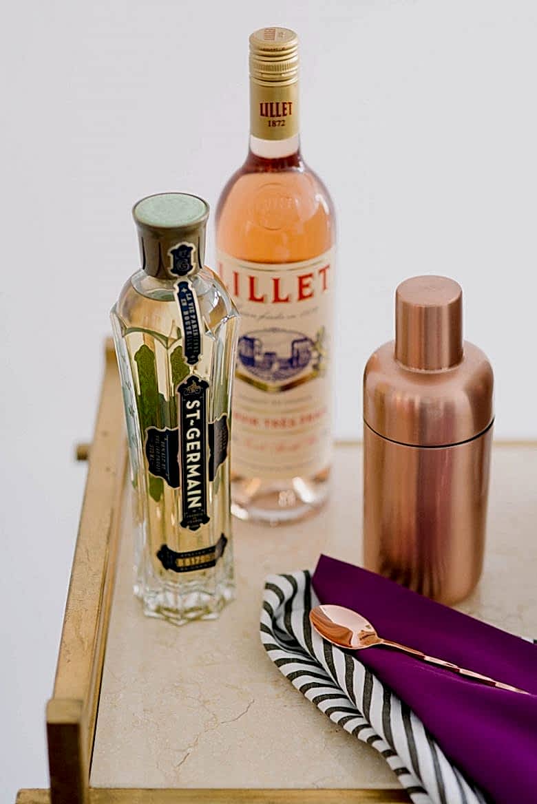 Bar cart detail of St. Germain, Lillet, a copper drinks shaker, and a copper bar spoon on purple and striped napkins