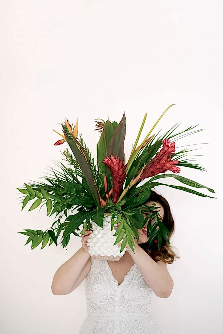 woman in white jumpsuit holding pineapple vase with tropical foliage