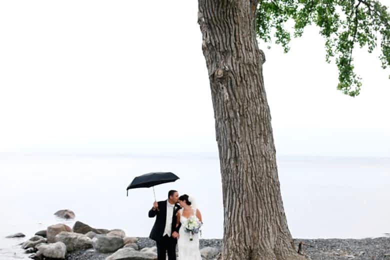 Newly wed couple under giant tree in front of water with large rocks, with groom holding black umbrella overhead