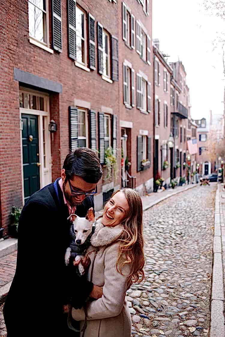 Smiling couple on cobblestone street with dog wearing a puffy vest