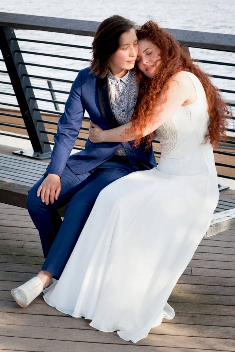 Couple in wedding attire embracing on bench near water