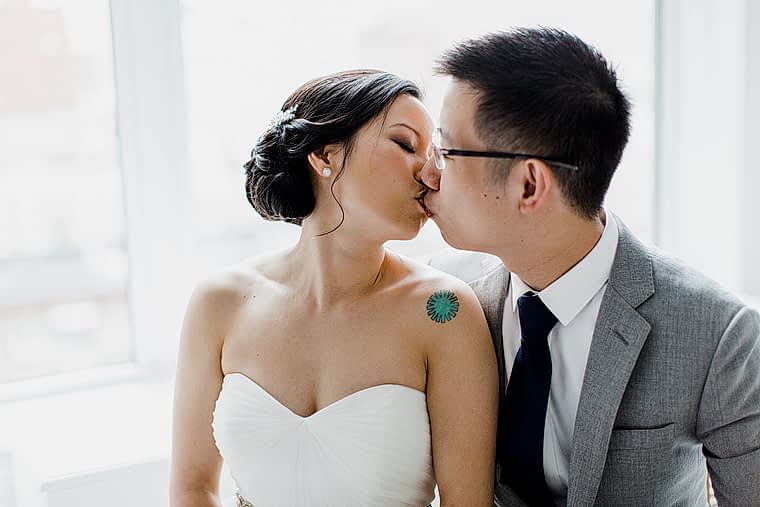 Bride with small blue shoulder tattoo kissing groom in grey suit near brightly lit window