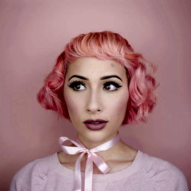 A woman with pink hair stands in front of a pink backdrop wearing a pink ribbon around her neck