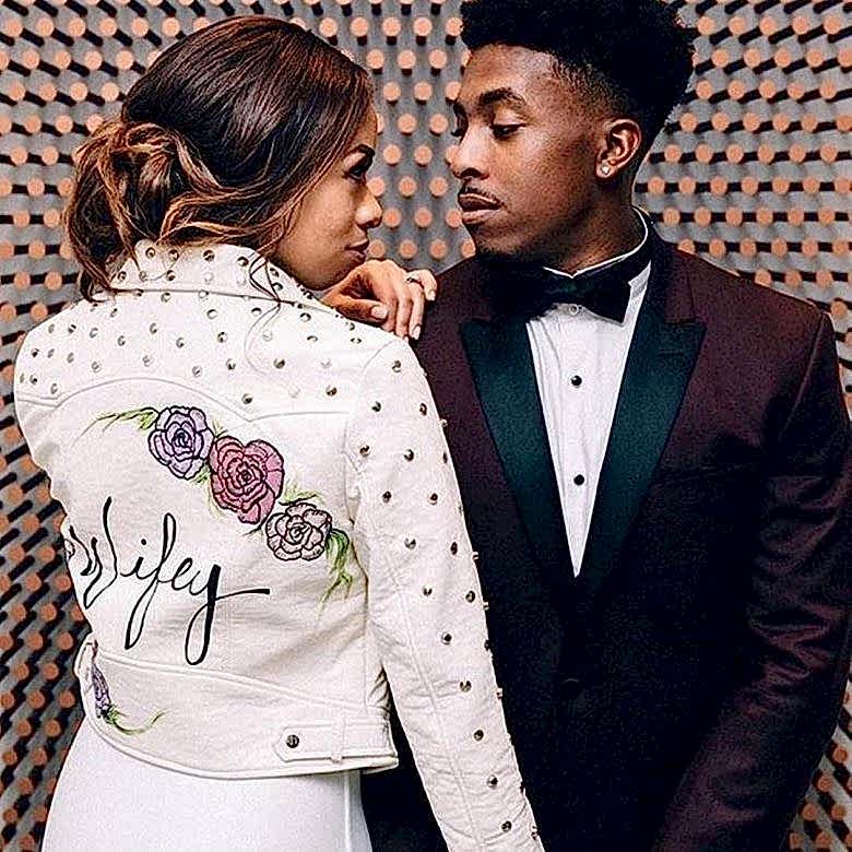 A bride and groom stand together with her back facing the viewer - she's wearing a white denim jacket with the word Wifey printed on the back