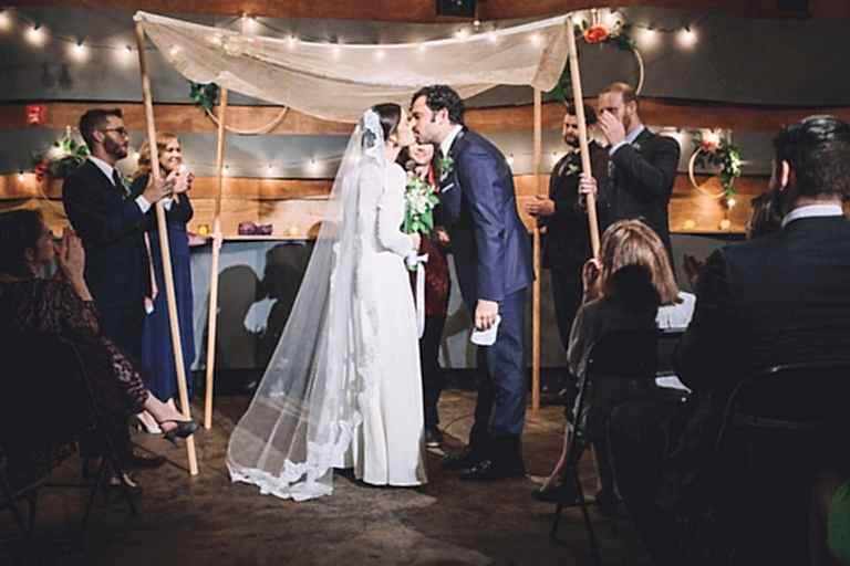 The bride & groom kiss during their ceremony, beneath the chuppah