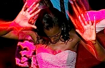 A bride bathed in neon pink light on the dance floor, dances with her face down and hands up mid-motion