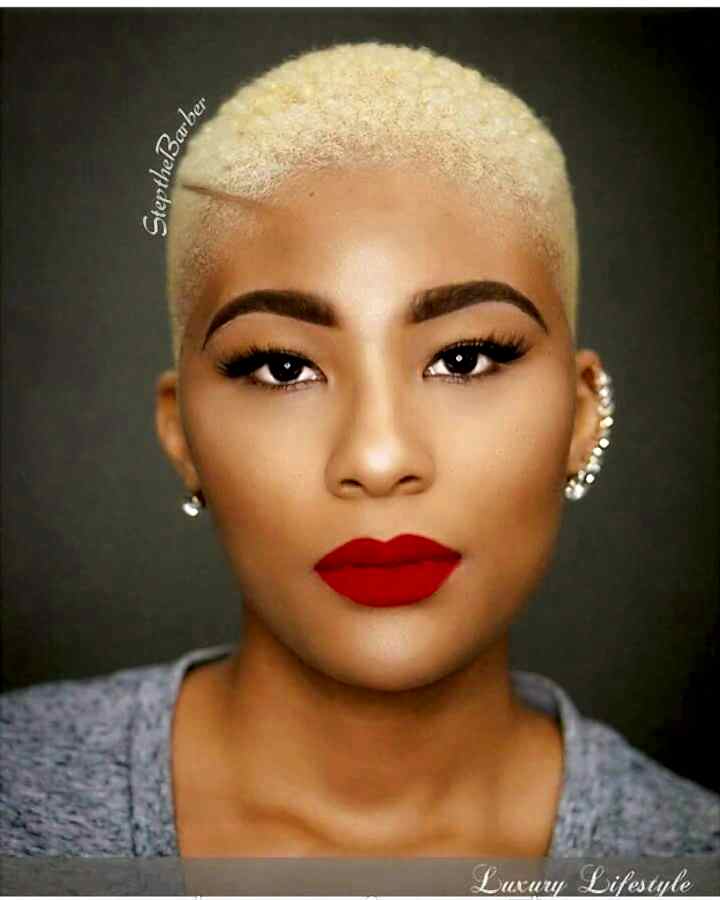 Portrait of woman with warm brown skin, bright red lips, bold dark eyebrows, and very close cropped blond hair