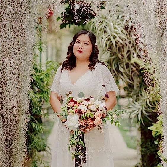 woman in garden beneath mossy curtain, holding pink and white bouquet, with medium length hair parted on side with loose waves