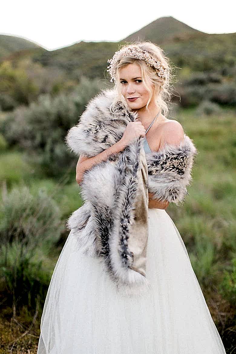 A bride stands in a grassy field wrapped in a luxurious faux fur shawl