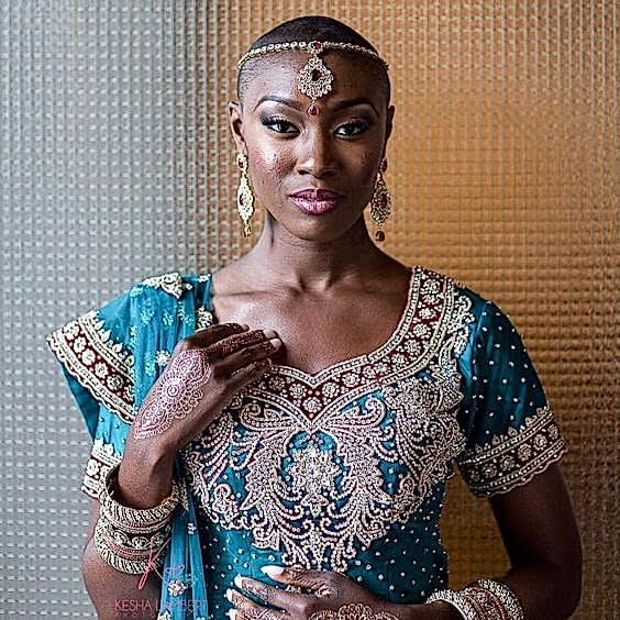 woman in ornate blue and red dress with gold appliqués, with close shaved dark hair, wearing a gold bejeweled headband with large red stone and gold tikka