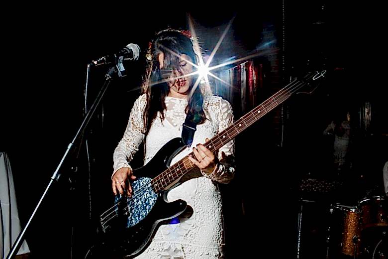 bride with long dark hair playing bass near microphone with light shining behind her
