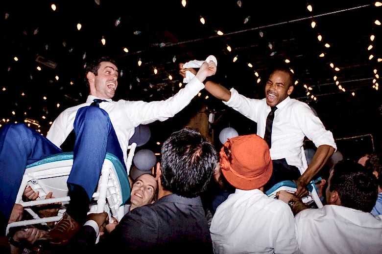 two joyful grooms being hoisted in chairs by wedding guests beneath illuminated string lights