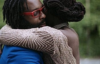 A man wearing a blue suit and red glasses holds a woman, her face looking away from the viewer.