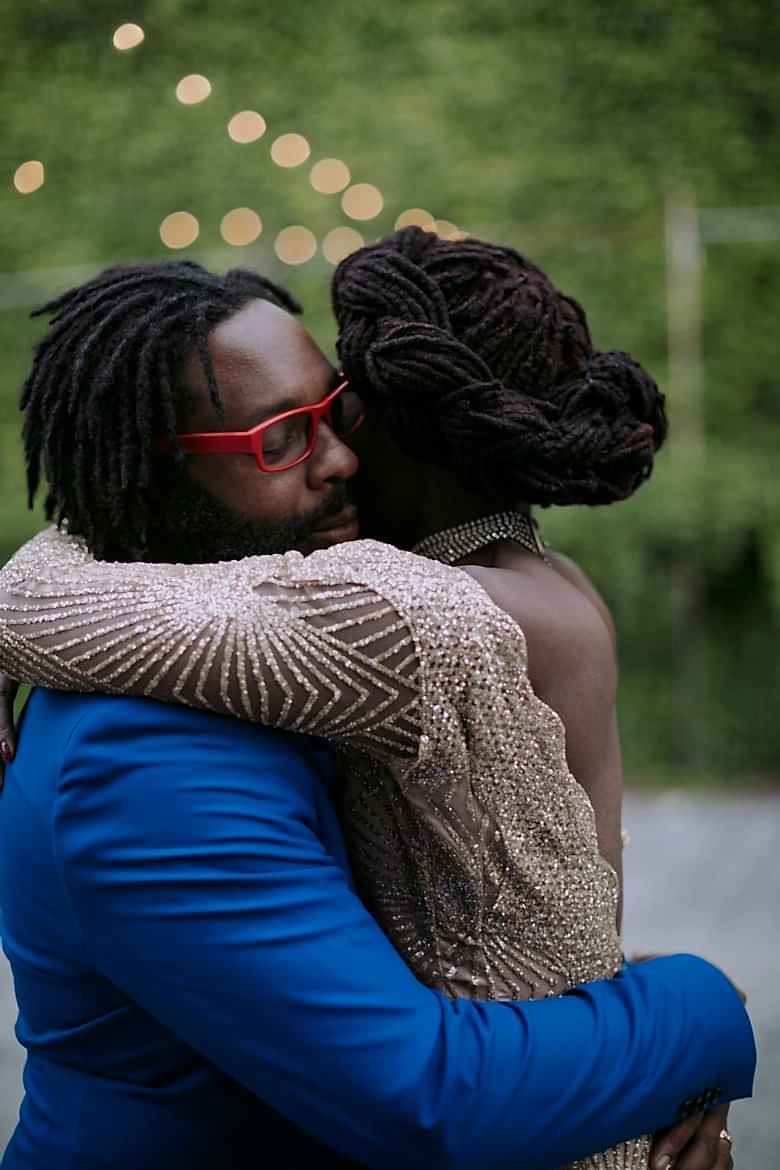 A man wearing a blue suit and red glasses holds a woman, her face looking away from the viewer.