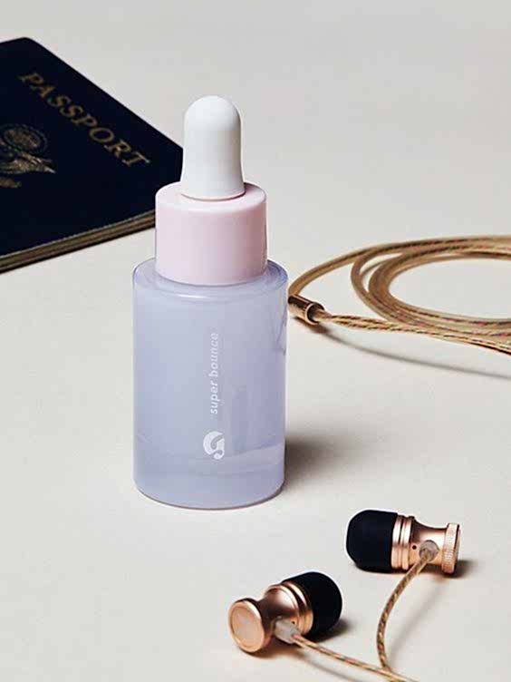 Bottle of Glossier Super Bounce serum on desk with headphones and passport in background