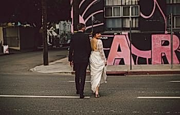 the wedding couple cross a city street as she looks over her shoulder back at the camera