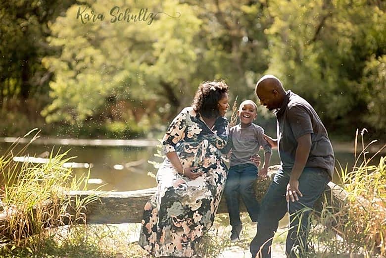 family sits on felled tree near lake, lauging in golden sunlight