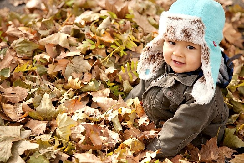 young child in a blue and fur flap cap sits in a pile of leaves, smiling