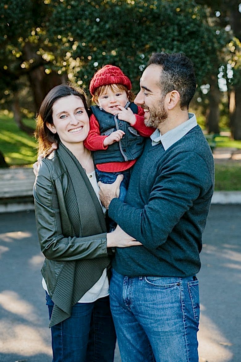 couple holding and hugging a young child in red hat in a park. they smile. 