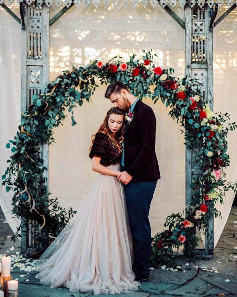 bride and groom in front of white wall, grey pillars, and large wreath of greenery, adorned on one side with red, pink, and white flowers, sitting on the ground 