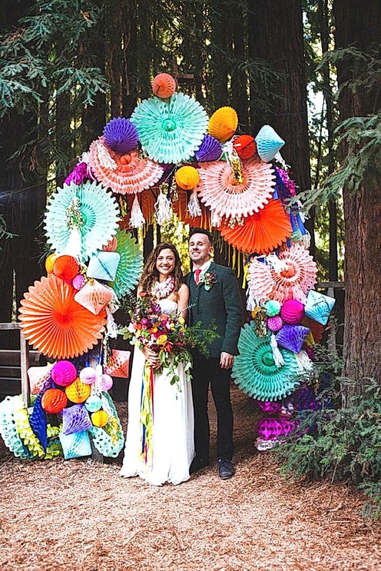 couple stands beneath an arch made of colorful paper pinwheels and honeycomb decorations
