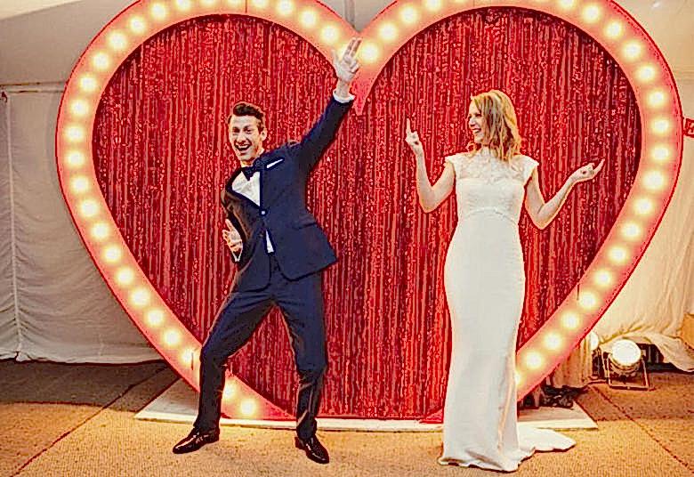 bride and groom stand in front of game-show style heart frame bordered in lights with sparkly red waterfall curtain filling 