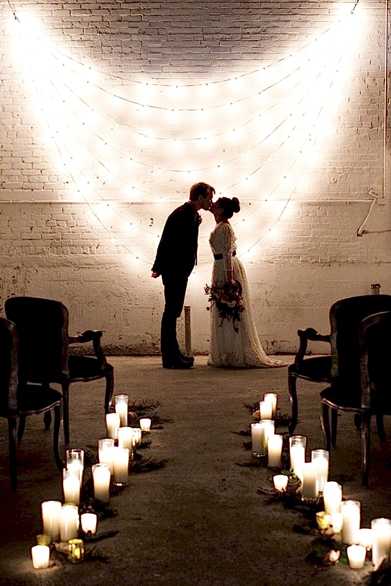 bride and groom kiss in front of a white brick wall with 7 strands of white string lights hung in a cascading scallop, aisle lined with white candles