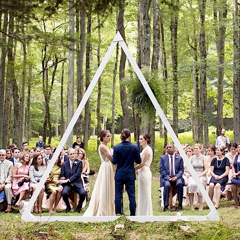 two brides with an officiant in front of guests at wedding ceremony in woods stand before a larg equilateral white triangle frame