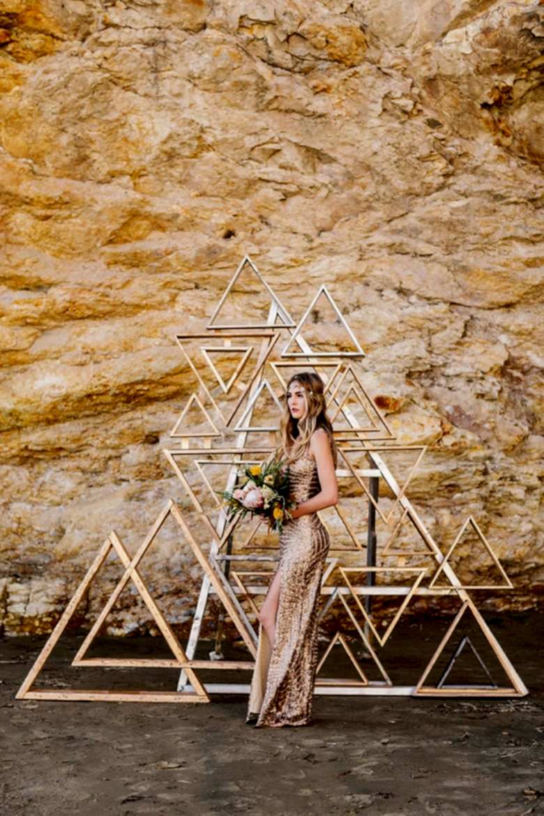 woman in sparkly gold cocktail-style dress stands in front of metallic triangle frame collage before a rock wall