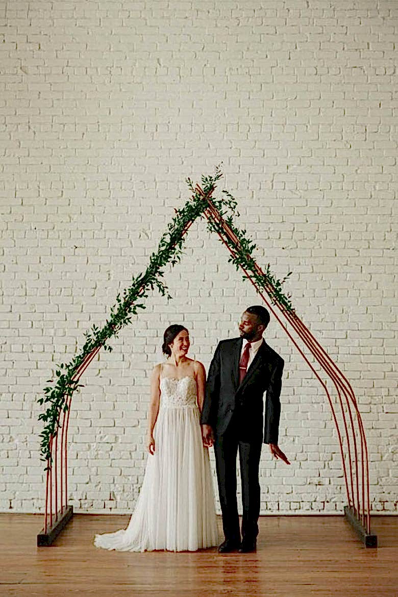bride and groom under minimal house-shaped arch with vine greenery coming up one side and halfway down the other, in front of a white brick wall