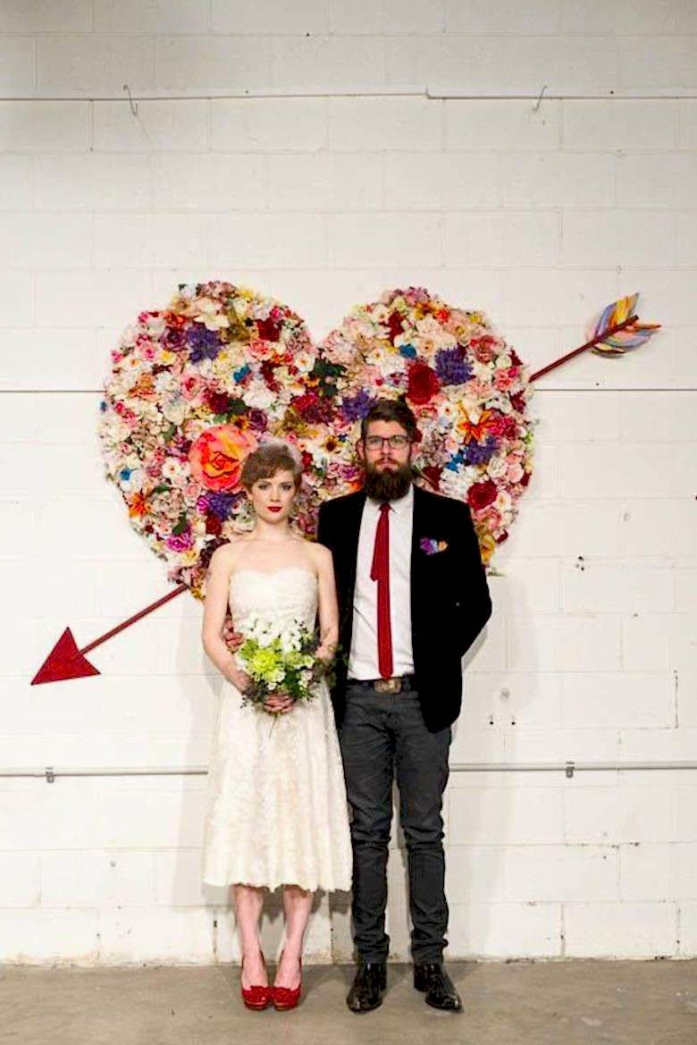 bride and groom stand in front of heart made of a flower collage, with an arrow through it