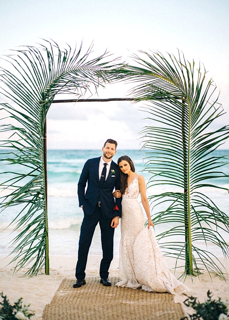 bride and groom at altar on beach framed by tall leafy fronds