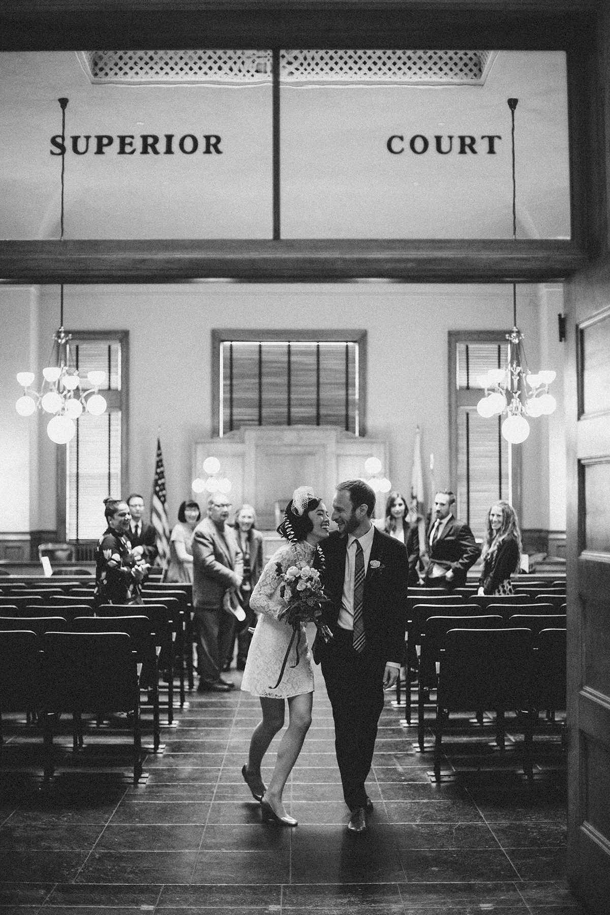 black and white photo of a bride in a short dress and groom during recessional down the aisle of a room that says "superior court" above the door in a Gina Clyne photo