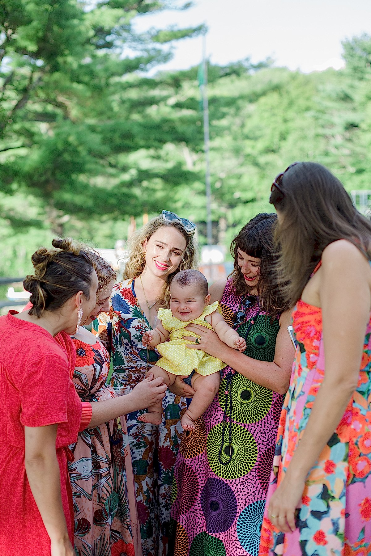 group of smiling women in brightly patterned dresses gather around one holding a baby in a Gina Clyne photo
