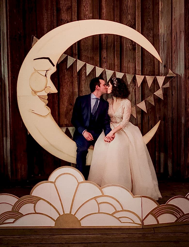 bride and groom sit kissing on hand-made paper moon in front of bunting with a deco-lights inspired 2-D cutout in front of them