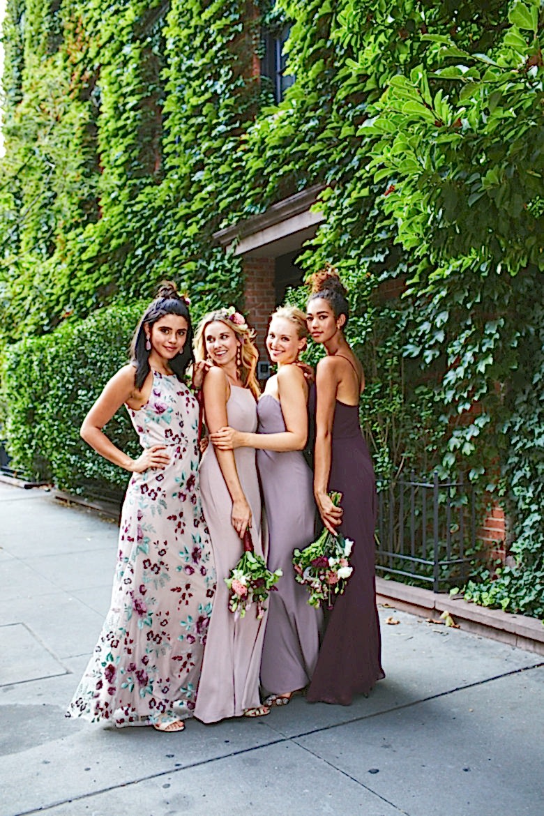 Group of bridesmaids wearing mismatched dresses in a floral and purple color palette against an ivy covered wall.